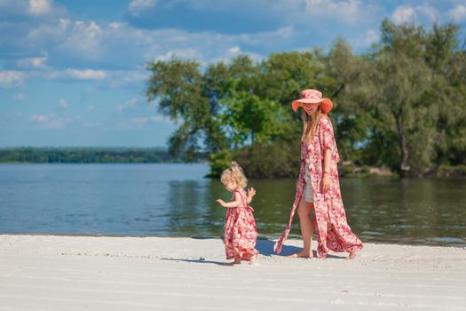 A charming girl in a light summer sundress walks on the sandy beach with her little daughter. Enjoys warm sunny summer days.