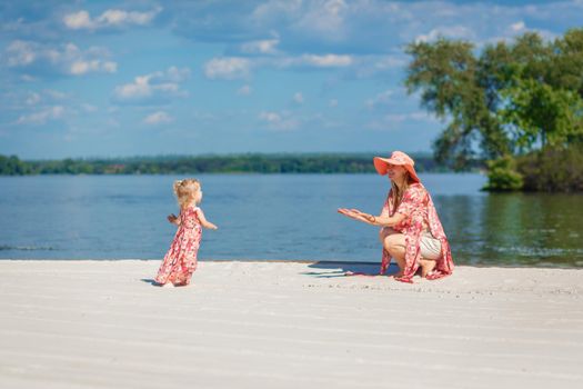 A charming girl in a light summer sundress walks on the sandy beach with her little daughter. Enjoys warm sunny summer days.