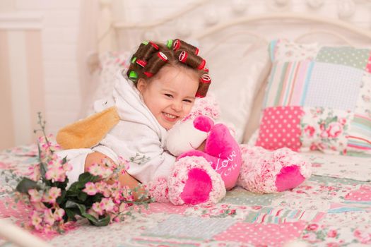 Adorable little girl with hair curlers hugs her favorite teddy bear toy.