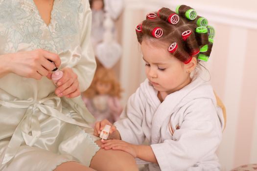 Adorable little girl with her mother in curlers paint their fingernails. Copies mom's behavior. Mom teaches her daughter to take care of herself. Beauty day.