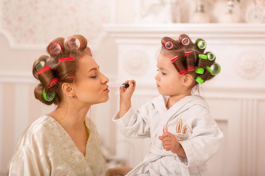 Adorable little girl with her mother in hair curlers apply makeup. Mom teaches daughter to use cosmetics. Beauty day. Girls are such girls.