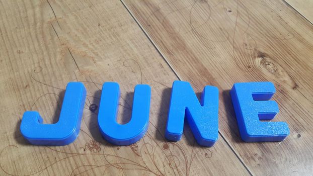 Plastic colored alphabets making word June are placed on a wooden floor. These plastic letters can be used for teaching kids.