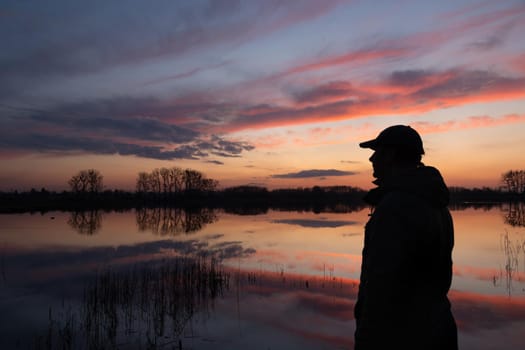 The silhouette of a man on the shore of a lake and sky by sunset, spring evening view