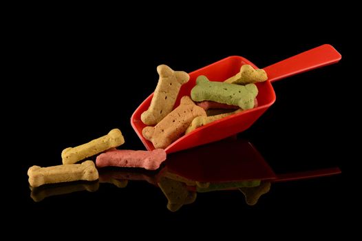 A red scoop of various dog treats in the shape of a bone over a black reflective surface.