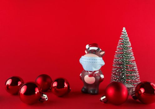Ceramic statuette of Ox in medical mask, Christmas tree and balls on a red backgroud with copy space. Symbol of New Year 2021.
