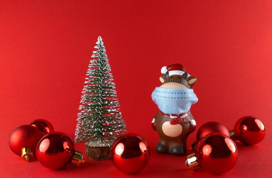 Ceramic statuette of Ox in medical mask, Christmas tree and balls on red backgroud. Symbol of New Year 2021.
