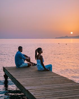 young romantic couple in love is sitting and hugging on wooden pier at the beach in sunrise time with golden sky. Vacation and travel concept. Romantic young couple dating at seaside. Crete Greece