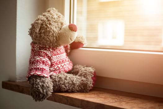 Cute teddy bear is sitting on the windowsill, looking out of the window, sunlight
