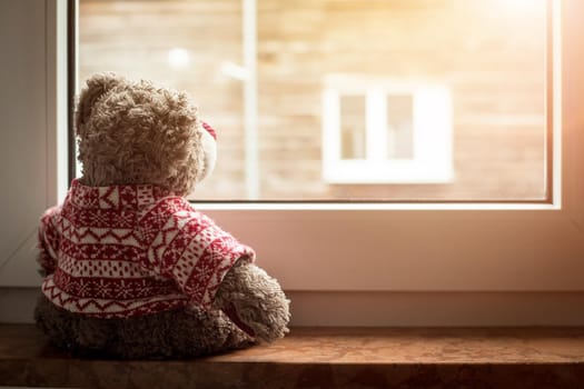 Cute teddy bear is sitting on the windowsill, looking out of the window, sunlight
