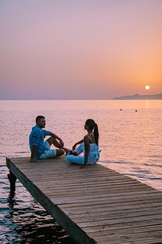 young romantic couple in love is sitting and hugging on wooden pier at the beach in sunrise time with golden sky. Vacation and travel concept. Romantic young couple dating at seaside. Crete Greece