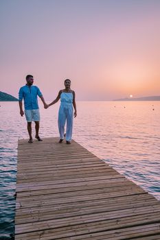 young romantic couple in love is sitting and hugging on wooden pier at the beach in sunrise time with golden sky. Vacation and travel concept. Romantic young couple dating at seaside. Crete Greece