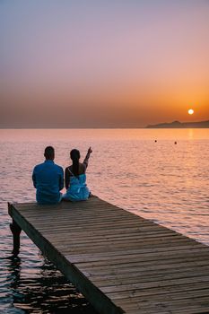 young romantic couple in love is sitting and hugging on wooden pier at the beach in sunrise time with golden sky. Vacation and travel concept. Romantic young couple dating at seaside. Crete Greece