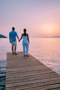 young romantic couple in love is sitting and hugging on wooden pier at the beach in sunrise time with golden sky. Vacation and travel concept. Romantic young couple dating at seaside. Crete Greece