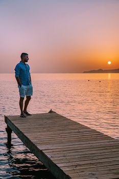 young romantic couple in love is sitting and hugging on wooden pier at the beach in sunrise time with golden sky. Vacation and travel concept. Romantic young couple dating at seaside. Crete Greece
