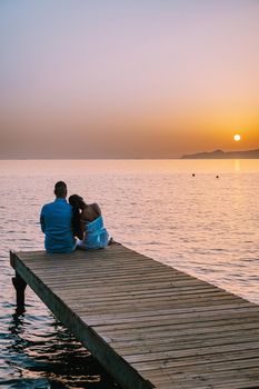 young romantic couple in love is sitting and hugging on wooden pier at the beach in sunrise time with golden sky. Vacation and travel concept. Romantic young couple dating at seaside. Crete Greece