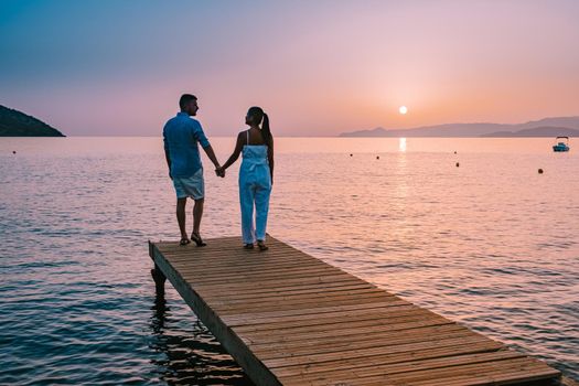 young romantic couple in love is sitting and hugging on wooden pier at the beach in sunrise time with golden sky. Vacation and travel concept. Romantic young couple dating at seaside. Crete Greece