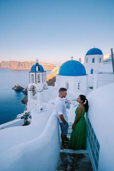 Santorini Greece, young couple on luxury vacation at the Island of Santorini watching sunrise by the blue dome church and whitewashed village of Oia Santorini Greece during sunrise during summer vacation, men and woman on holiday in Greece