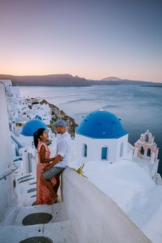 Santorini Greece, young couple on luxury vacation at the Island of Santorini watching sunrise by the blue dome church and whitewashed village of Oia Santorini Greece during sunrise during summer vacation, men and woman on holiday in Greece