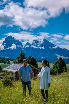 Alpe di Siusi - Seiser Alm with Sassolungo - Langkofel mountain group in background at sunset. Yellow spring flowers and wooden chalets in Dolomites, Trentino Alto Adige, South Tyrol, Italy, Europe. Summer weather with dark clouds rain, couple men and woman on vacation in the Dolomites Italy