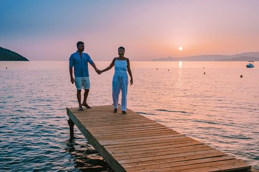 young romantic couple in love is sitting and hugging on wooden pier at the beach in sunrise time with golden sky. Vacation and travel concept. Romantic young couple dating at seaside. Crete Greece