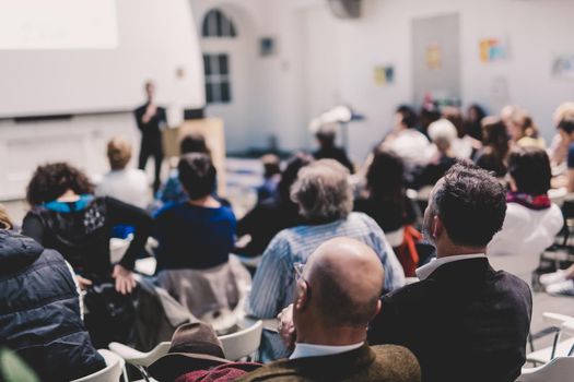 Business and entrepreneurship symposium. Speaker giving a talk at business meeting. Audience in the conference hall. Rear view of unrecognized participant in audience.