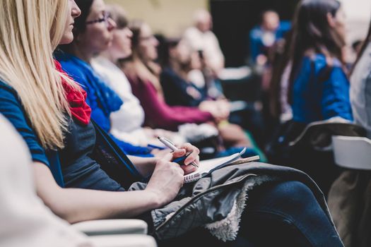 Female hands holding pens, notebook and phone and making notes at conference lecture. Event participants in conference hall.