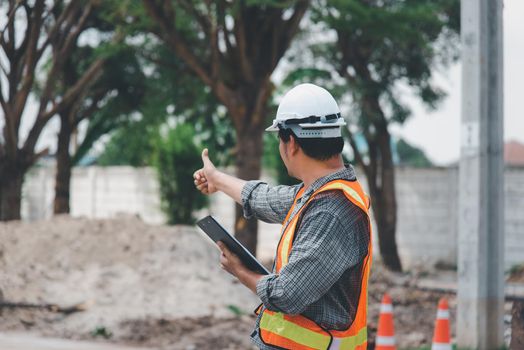 Asian man civil construction engineer worker or architect with helmet and safety vest working and holding a touchless tablet computer for see blueprints or plan at a building or construction site