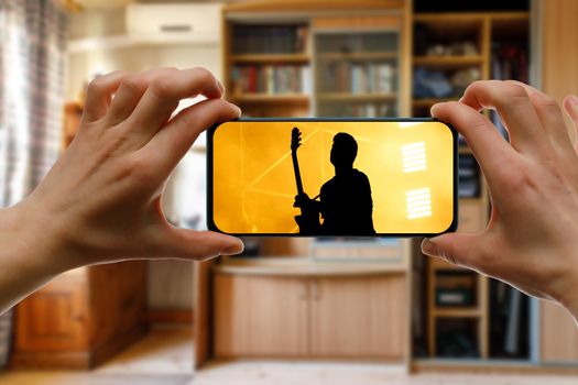 Watching online music concert at home using a mobile phone. Guitarist silhouette on the screen