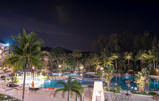 Swimming pool area in a beach hotel resort during night with trees and smooth blue swimming pool water reflecting lights long exposure somewhere in Asia