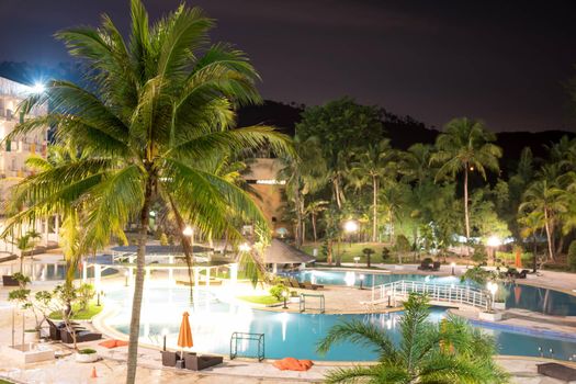 Swimming pool area in a beach hotel resort during night with trees and smooth blue swimming pool water reflecting lights long exposure somewhere in Asia