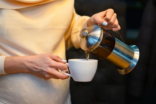 hands of a pregnant girl in a light white and yellow jacket, who hold a mug in which she pours steamed tea from a teapot