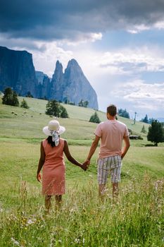 Alpe di Siusi - Seiser Alm with Sassolungo - Langkofel mountain group in background at sunset. Yellow spring flowers and wooden chalets in Dolomites, Trentino Alto Adige, South Tyrol, Italy, Europe. Summer weather with dark clouds rain, couple men and woman on vacation in the Dolomites Italy