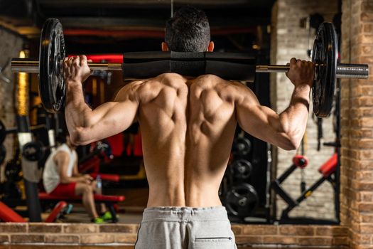 Back view of young bodybuilder flexing muscles with barbell in front of mirror at gym