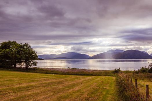 Beautiful mystic landscape lake scenery in Scotland with cloudy sky, meadow, trees and sunbeams