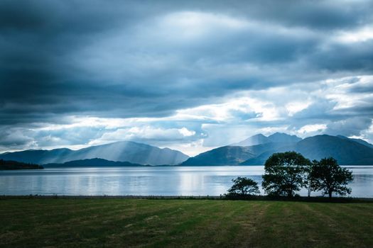 Beautiful mystic landscape lake scenery in Scotland with cloudy sky, meadow, trees and sunbeams