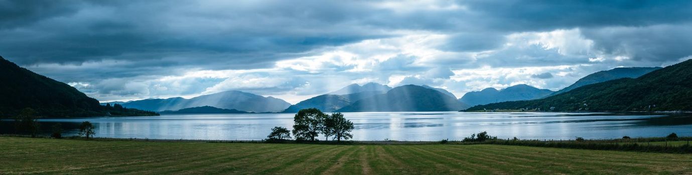 Beautiful mystic landscape lake scenery in Scotland with cloudy sky, meadow, trees and sunbeams