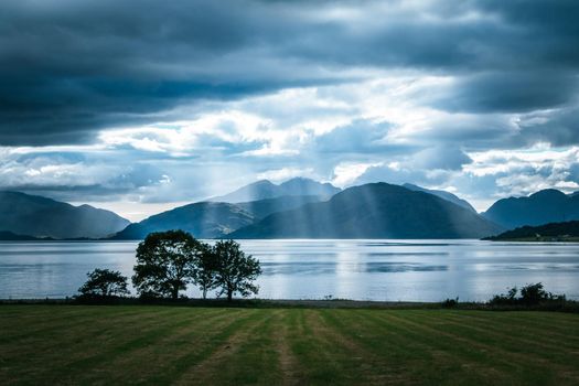 Beautiful mystic landscape lake scenery in Scotland with cloudy sky, meadow, trees and sunbeams