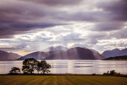 Beautiful mystic landscape lake scenery in Scotland with cloudy sky, meadow, trees and sunbeams