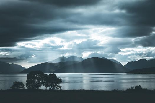 Beautiful mystic landscape lake scenery in Scotland with cloudy sky, meadow, trees and sunbeams