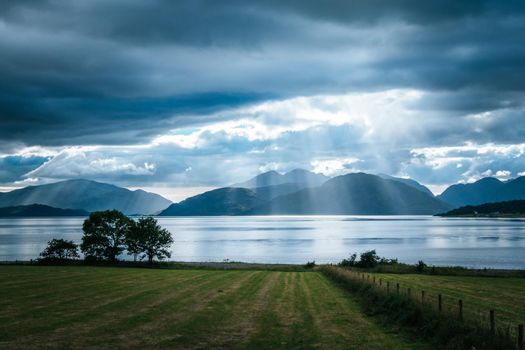 Beautiful mystic landscape lake scenery in Scotland with cloudy sky, meadow, trees and sunbeams