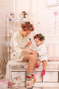 Adorable little girl with her mother in curlers paint their fingernails. Copies mom's behavior. Mom teaches her daughter to take care of herself. Beauty day.