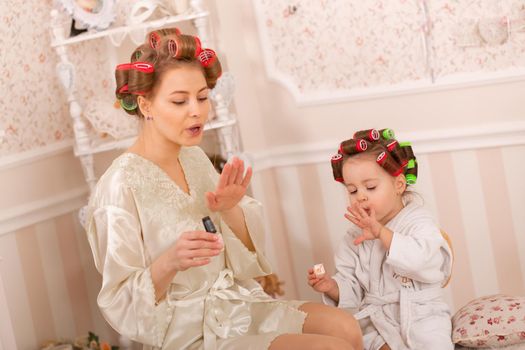 Adorable little girl with her mother in curlers paint their fingernails. Copies mom's behavior. Mom teaches her daughter to take care of herself. Beauty day.