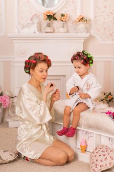 Adorable little girl with her mother in hair curlers apply makeup. Mom teaches daughter to use cosmetics. Beauty day. Girls are such girls.