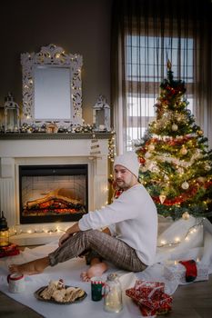Cute, young guy mid age by fireplace with a Christmas tree,Family sitting on a floor. men near christmas tree with candle lights