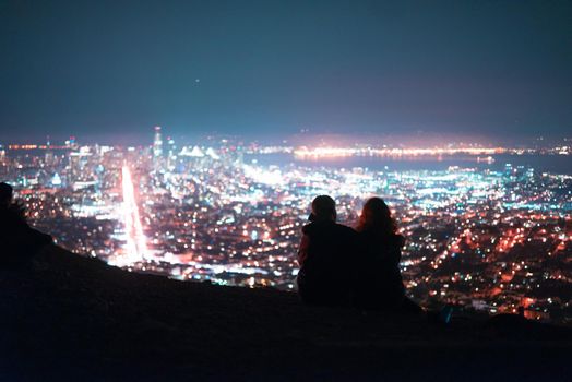 A couple sits on a hill looking out over the city lights of San Francisco.