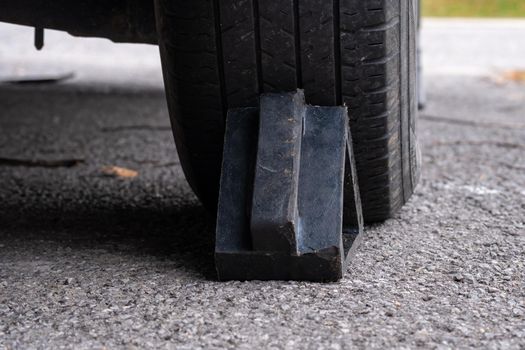 A black rubber wheel chock is seen from behind as it braces a rear, passenger-side tire on a car. It sits on a paved driveway.