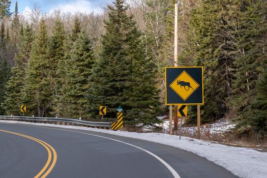 A large moose crossing sign with a moose icon inside a yellow diamond shape warns motorists of the hazard of crossing moose. The warning stands on the side of a road in early winter, with light snow.
