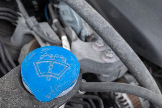 A blue cap for a windshield washer fluid storage tank is seen under the hood of a car. The cap and surrounding mechanical components are dirty.