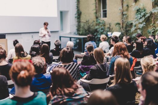 Female speaker giving presentation in lecture hall at university workshop. Audience in the conference hall. Rear view of unrecognized participant in audience. Scientific conference event.