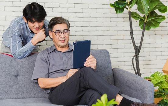 Asian senior businessman with digital tablet discuss together with young team in office. Father man and his son sit on sofa talking chatting on video call conference on tablet in living room at home